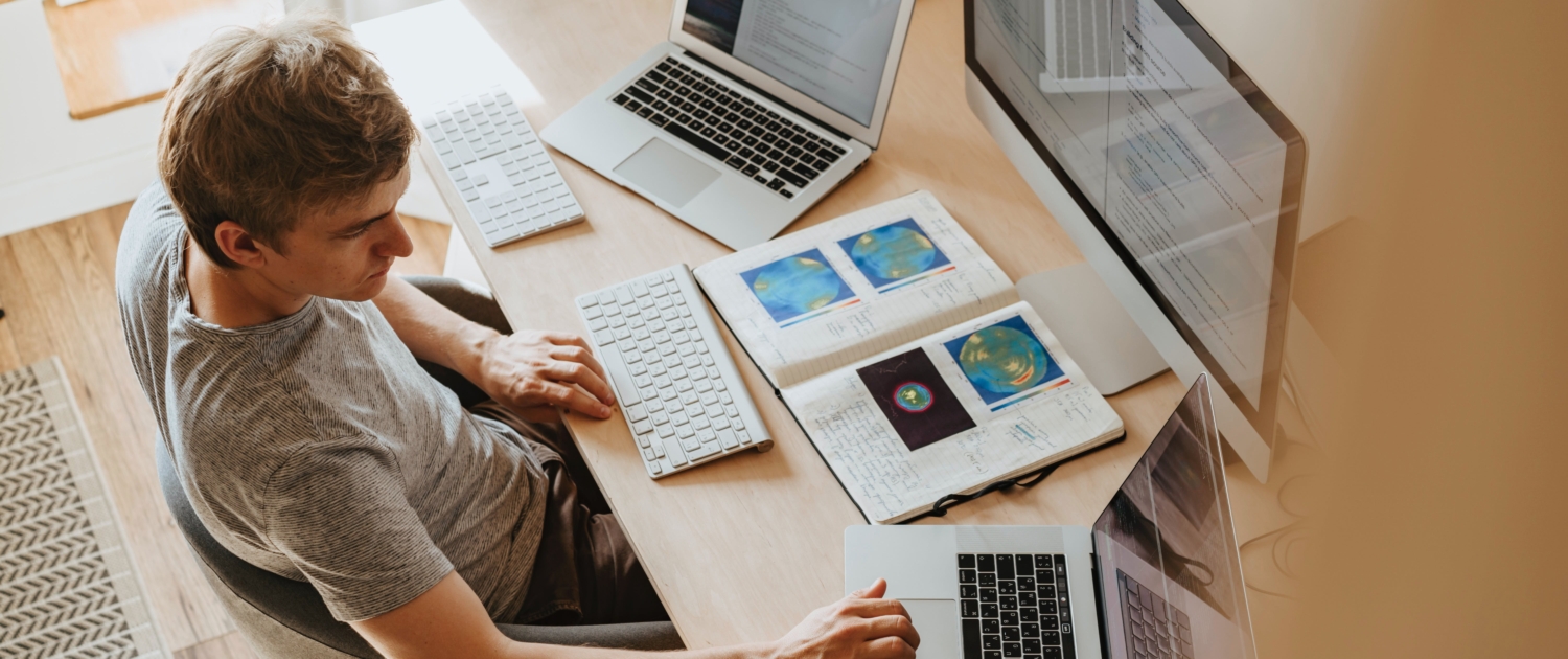 man using multiple computers at once, reports scattered on his desk.