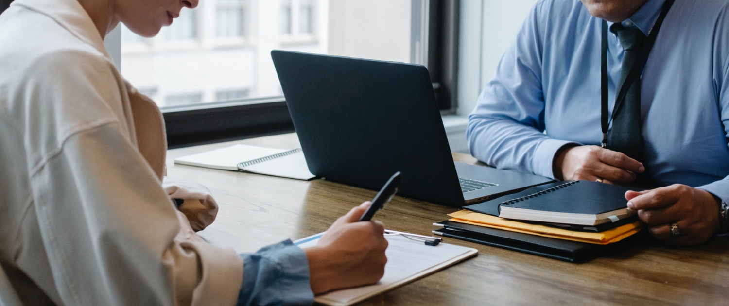 Two people in an office setting, one on a laptop the other writing on a paper.